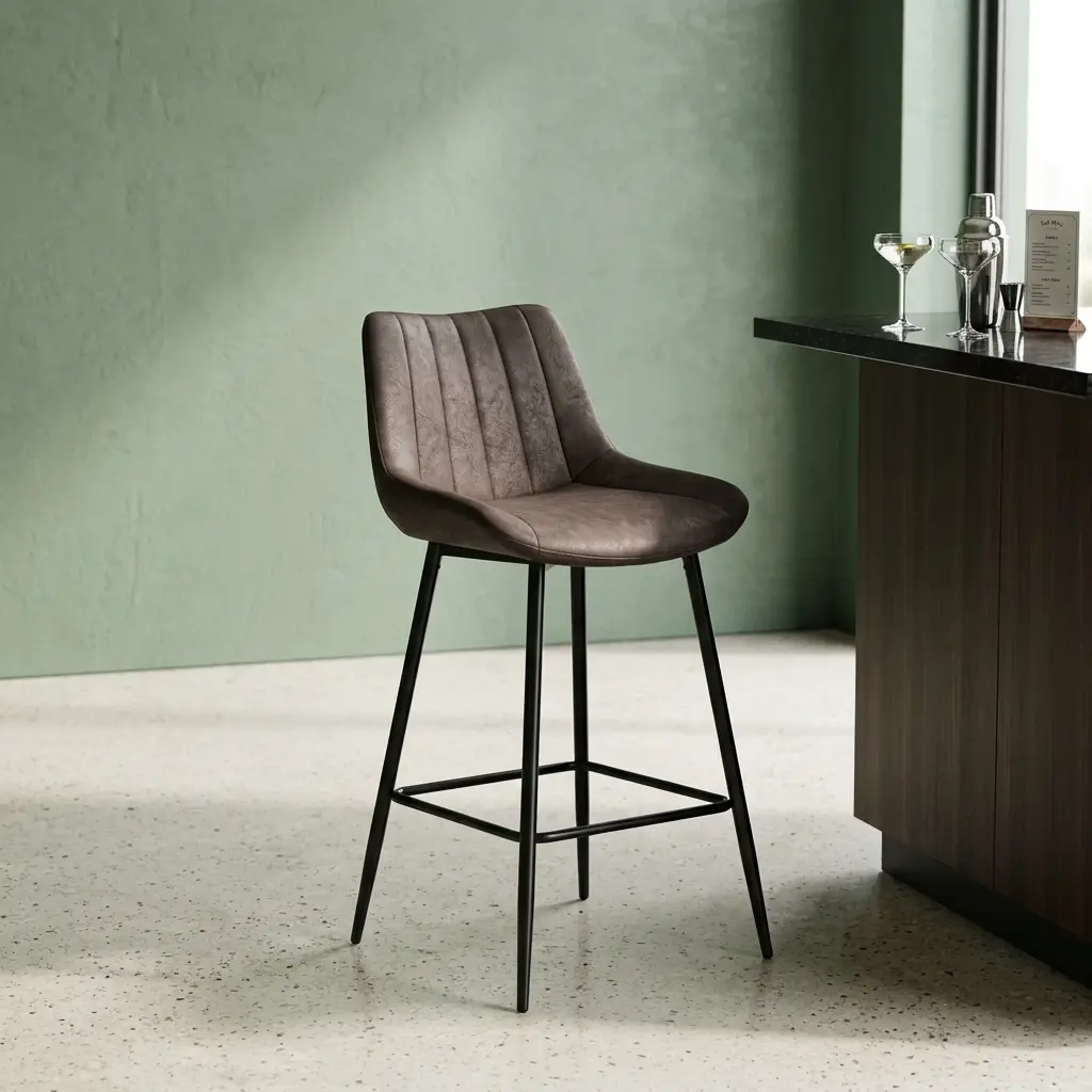 Dark gray leatherette kitchen barstool with vertical channel-tufted low backrest, bucket seat design, black tapered metal legs, and integrated square footrest.