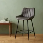 Dark gray leatherette kitchen barstool with vertical channel-tufted low backrest, bucket seat design, black tapered metal legs, and integrated square footrest.
